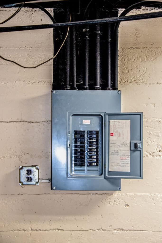 Stainless steel electrical circuit box open with circuits named by hand in basement on rough concrete block wall with conduit and ceiling and other pipes overhead painted black with dust on them